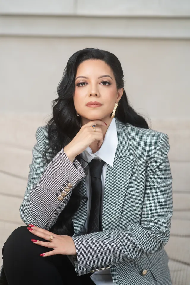 Priscilla Laclé in a checked blazer and tie, resting her chin on her hand