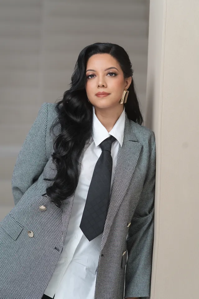 Priscilla Laclé leaning against a wall in a checked blazer